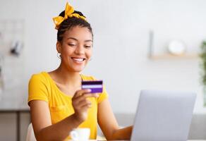 Online Wallet And Banking Concept. Smiling black woman using laptop and credit card, making financial transaction, sitting at desk, ordering purchases, checking debit card number and security code photo