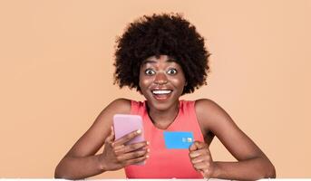 Mobile Shopping Application. Excited Black Lady Using Phone And Credit Card For Shopping Posing With Clothing Rack Over Yellow Studio Background, Smiling To Camera. photo