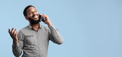 Modern Communication Concept. Smiling African American Bearded Man Talking On Cellphone Standing Over Blue Studio Background, Isolated. Cheerful Black Guy Using Gadget To Make Phone Call, Gesturing photo