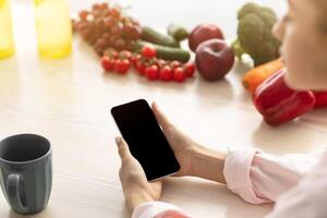 Search Recipe. Over the shoulder view of young lady holding and using smartphone with blank black empty screen for mock up design. Female sitting at dinner table with cup of coffee photo