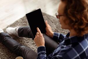 Unrecognizable Man Using Digital Tablet With Blank Screen For Application Or Website Advertisement. Back View Of Guy Browsing Internet On Computer Sitting On Floor At Home. Mockup, High Angle photo