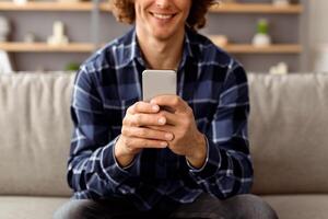 Unrecognizable Hipster Guy Using Mobile Phone Texting And Browsing Internet, Networking In Social Media Online Sitting On Sofa Holding Smartphone At Home. Man With Cellphone Using Application. Cropped photo