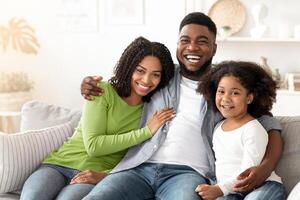 Family lifestyle portrait of happy black mom, dad with their little daughter at home, hugging on couch in living room and looking at camera photo