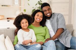 Happy family portrait. Loving black father, mother and little daughter hugging on couch and smiling to camera, enjoying staying at home, free space photo
