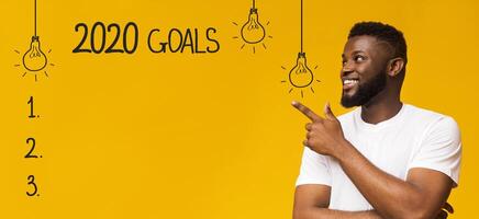 Future plans. Cheerful black man pointing at checklist with 2020 goals, yellow background photo