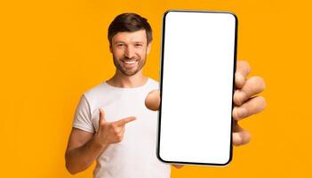Recommendation. Portrait of excited bearded man holding big smartphone with white blank screen in hand, showing close to camera and pointing at device. Gadget with empty free space for mock up, banner photo