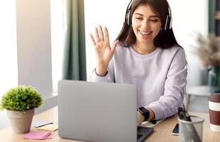 Online Meeting Concept. Young smiling woman making call on laptop at home, positive female in wireless headset waving at computer web camera, enjoying distance communication, copy space photo