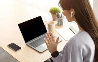 Modern Remote Communication Concept. Over the shoulder view of young woman making online call using laptop and waving to blank empty screen for mock up, sitting at desk wearing earphones photo