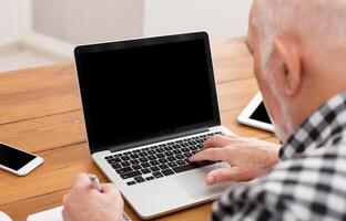 Senior man using laptop with blank screen back view, mockup. Mature male making call with computer at home, copy space photo