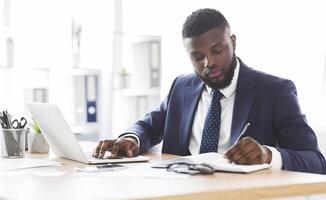 Successful american businessman working hard in office, making notes in front of laptop, panorama with free space photo