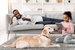 Weekend Concept. Black man lying on couch, watching television, holding remote control, happy puppy lying on carpet, smiling girl sitting on floor using tablet spending time together. Family Life photo