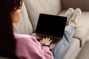 People And Technology Concept. Over the shoulder view of woman using laptop with black empty screen for mockup template, typing on keyboard, sitting on couch, studying or working online at home photo