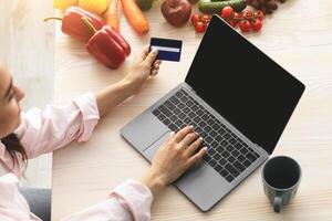 Easy Payment. Above top high angle view of smiling young woman using laptop sitting at dinner table, holding and showing debit credit card. Housewife ordering food, free copy space photo