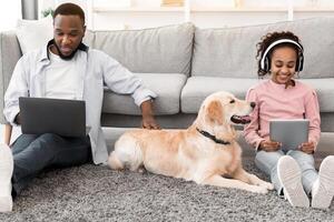 Leisure Time. African American girl sitting on rug floor carpet with dog and holding digital tablet, wearing headphones, man using computer and working at home office browsing internet, patting dog photo