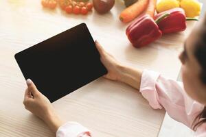 Over the shoulder view of young woman holding and using digital tablet with black empty screen for mock up template, cooking in kitchen, drinking coffee in the morning, searching new recipes online photo