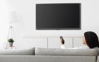 African american woman watching TV pointing remote control at flatscreen plasma television set with blank screen, switching channels while sitting on sofa at home, mockup photo