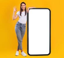 Happy Woman Leaning On Big Smartphone With Blank White Screen And Gesturing Ok Sign, Cheerful Lady Recommending New App Or Website, Standing On Yellow Background, Mock Up Image, Full Body Length photo