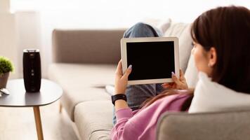 Over The Shoulder View Of Unrecognizable Young Woman Holding And Using Digital Tablet With Empty Mockup Screen, Sitting On Couch In Living Room At Home. Selective Focus On Gadget, Template photo