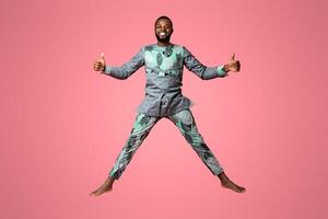 Happy black guy in traditional grey clothes jumping up over pink studio background, full length photo. Joyful african american man having fun while posing at studio, showing thumbs up, copy space photo
