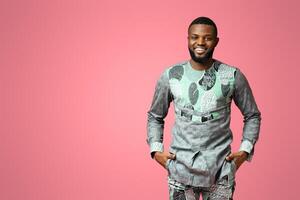 Confident black man in traditional african costume smiling on pink studio background, putting hands in pockets, copy space. Cheerful african american guy posing in ethnic colorful clothes photo