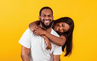 Love And Care. Portrait of smiling African American woman hugging her man from the back, enjoying time together, standing isolated over yellow studio background, posing and looking at camera photo