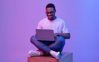 Handsome Black Programmer Guy Using Laptop Computer, Sitting On Big Cube In Neon Lighting, Cheerful African American Man In Glasses Working Or Study Online Over Purple Studio Background, Copy Space photo
