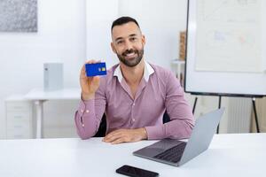A man with a beard smiles while displaying a blue card in his hand. He is seated at a desk with a laptop and a smartphone, suggesting a professional setting and an engaging moment. photo