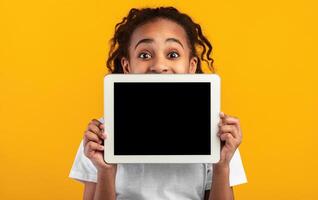 Great Application. Excited Black Girl Holding And Showing Digital Tablet With Empty Screen For Mockup, Covering Face And Mouth, Recommending New App Posing Standing Over Yellow Studio Background photo