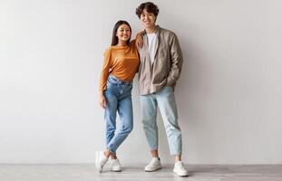 Full length portrait of happy young Asian couple in casual clothes hugging and looking at camera against white studio wall. Cheerful millennial spouses posing and smiling together photo