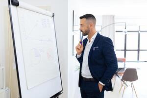 A well-dressed man stands pensively by a flip chart filled with graphs, engaged in a strategic discussion in a modern office setting during the day. photo
