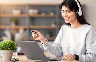 People And Modern Technology Concept. Portrait of smiling woman in wireless headset sitting at desk, using laptop and writing in notebook, taking notes, watching lecture or working, holding pen photo