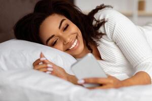 Portrait of happy African American woman lying in bed and smiling while reading text message from someone, holding and using smartphone, chatting with lover or browsing social media photo