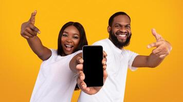 Advertising New Mobile App. Portrait of happy black couple holding smartphone with empty screen in hands, showing and pointing at device with blank space for mockup, yellow studio background photo