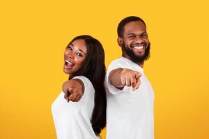 Your Turn. Portrait Of Excited African American Young Couple Pointing Fingers At Camera, Happy Man And Woman Choosing You, Standing Back To Back Isolated Over Yellow Studio Background, Copy Space photo