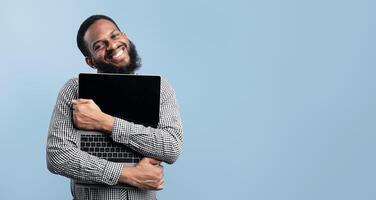 Funny crazy african guy embracing personal computer with empty display, isolated over turquoise studio background photo