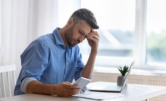 Worried male freelancer looking at smartphone screen while working at home office, received bad news, got scam message, thoughtful man touching forehead with concerned face expression, copy space photo