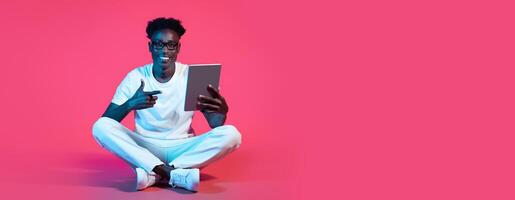 A young black man sits on the floor with his legs crossed, smiling as he interacts with a tablet. He wears a white t-shirt and white pants, creating a striking contrast with the pink backdrop. photo