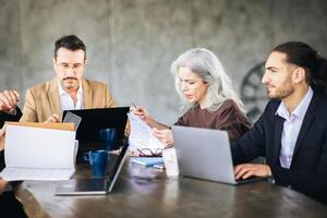 A diverse group of professionals engage in a brainstorming session around a table, using laptops and discussing ideas in a contemporary office environment. photo