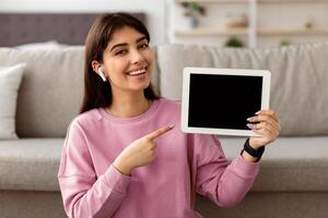 Check This Out. Portrait of smiling female person in earbuds holding and showing digital tablet with black empty screen in hand, presenting and pointing at gadget screen with blank space for mock up photo