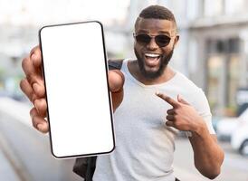 Great Offer. Portrait of excited black man standing outdoors holding big phone with white blank screen in hand, showing close to camera pointing at device. Gadget with empty free space for mock up photo