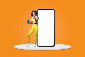 A fit woman stands confidently next to a large smartphone, holding a refreshing health drink. She wears bright yellow activewear, radiating energy and vitality in a vibrant setting. photo