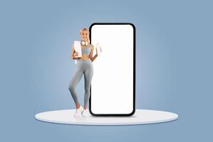 A fitness enthusiast stands confidently while holding a notebook and a drink. She is positioned next to a large blank smartphone screen, set against a light blue background. photo