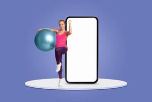 A woman in activewear confidently demonstrates an exercise routine using a fitness ball, while a large blank smartphone screen serves as a backdrop, creating an energetic atmosphere. photo
