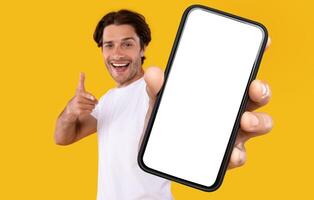 Cheerful young guy holding smartphone with white empty screen, pointing at blank display for mobile application advertisement or your text, gadget mockup template, isolated orange studio background photo