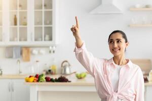 Modern Futuristic House Control System, IoT Technologies Concept. Portrait Of Smiling Millennial Lady Pointing Finger Up In The Air Pushing Button On Invisible Screen Standing In Kitchen Interior photo