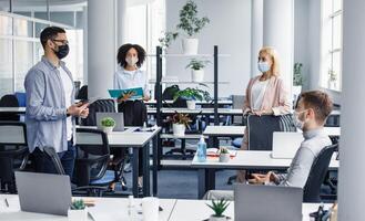Meeting with manager and protecting health after returning to office after quarantine. Boss in protective mask with tablet speaks to multiracial of workers in office interior with devices, empty space photo