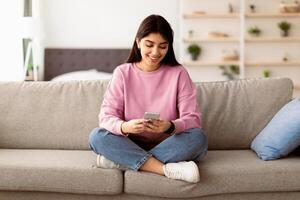 Cool Gadget And Application. Portrait of young smiling woman holding mobile phone, typing sms message, sitting on the sofa in living room. Woman browsing internet, surfing web, using app photo