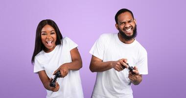 Couple Pastime Concept. Emotional black guy and lady playing online games, using joystick gamepad. Millennial family of two enjoying games, standing at blue studio, frontal portrait photo