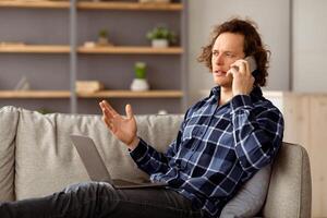 Serious Freelancer Guy Talking On Cellphone Sitting With Laptop On Couch, Communicating Working Online From Home In Living Room. Mobile Communication, Business Call Concept photo