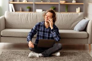 Mobile Communication. Cheerful Guy Talking On Smartphone Sitting With Laptop On Floor At Home, Working Remotely Online. Man Calling Speaking By Phone Having Pleasant Conversation photo